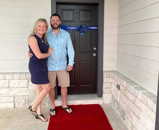 A couple standing in front of the front door of their new David Weekley Home. A red carpet leads to the front door, which is decorated with a blue, David Weekley Homes-branded bow and ribbon.