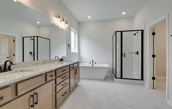 A bright interior photograph of a en suite bathroom in a new home with white quartz countertops at the double-sink vanity, light wood cabinets, white tile floors, a free-standing tub, glass shower enclosure, and modern rustic hardware.