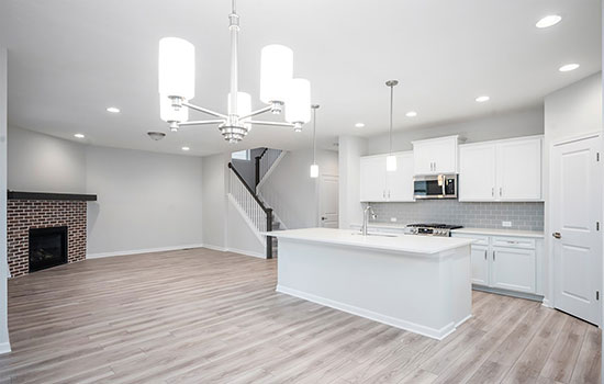 A bright interior photograph of a new home with a white interior paint, wood tile floors, a red brick fireplace and stainless steel appliances and fixtures in the kitchen. A staircase leading to the second floor can be seen beyond the family room, dining space and kitchen.