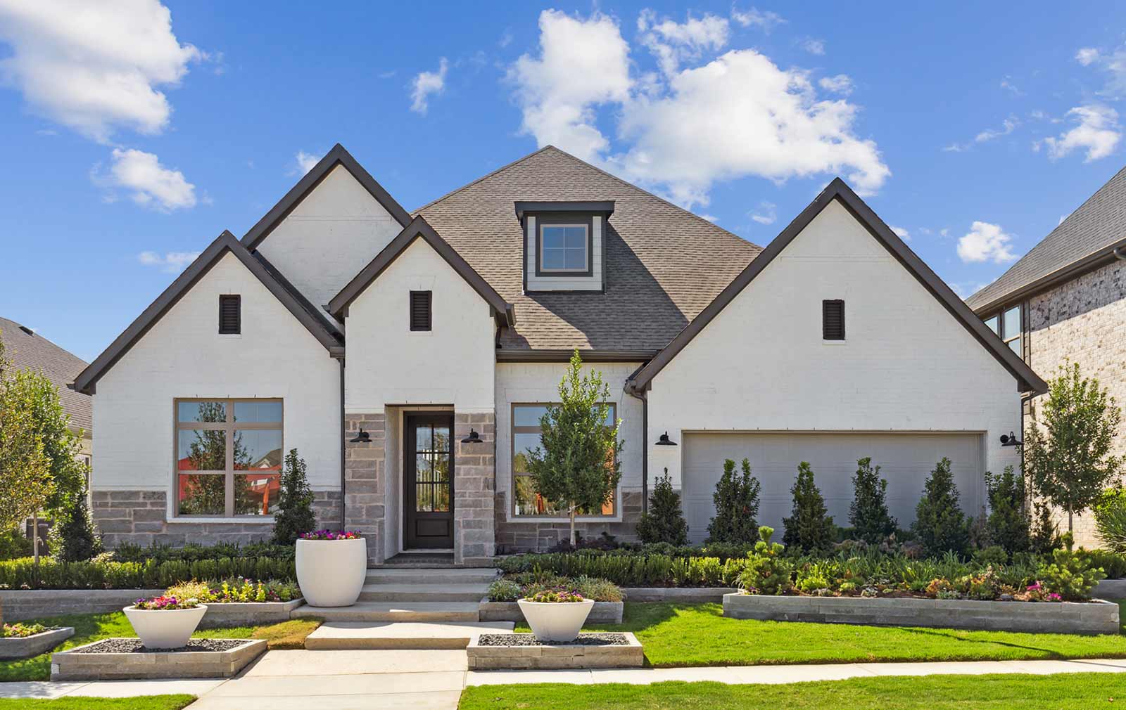 The front of a new construct model home with a cut stone and white brick exterior, multi-level landscaping, and puffy clouds dotting the clear blue sky in the background.