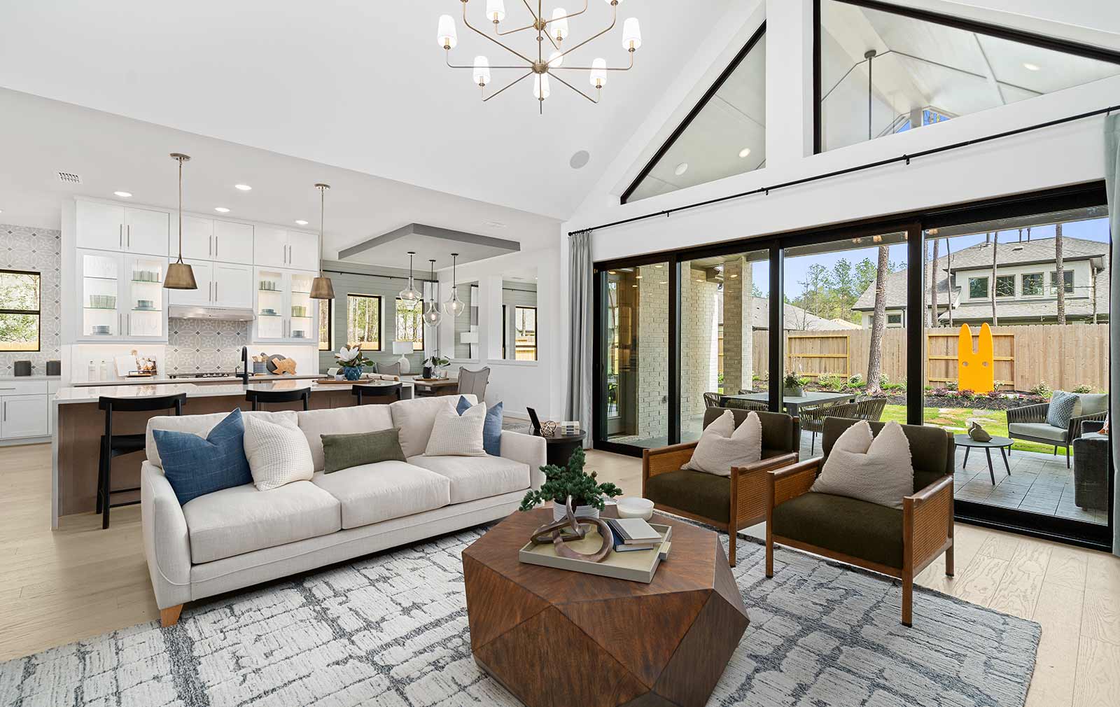 A model home’s interior, with the family room in the foreground with green, soft white and wood-tone furniture, an expansive kitchen and dining space in the background to the left, and a view of the back garden through the big, floor-to-ceiling windows to the right. The shape of a big yellow rabbit sculpture in the garden is mirror in the set of the throw pillows on the interior sofa and chairs.