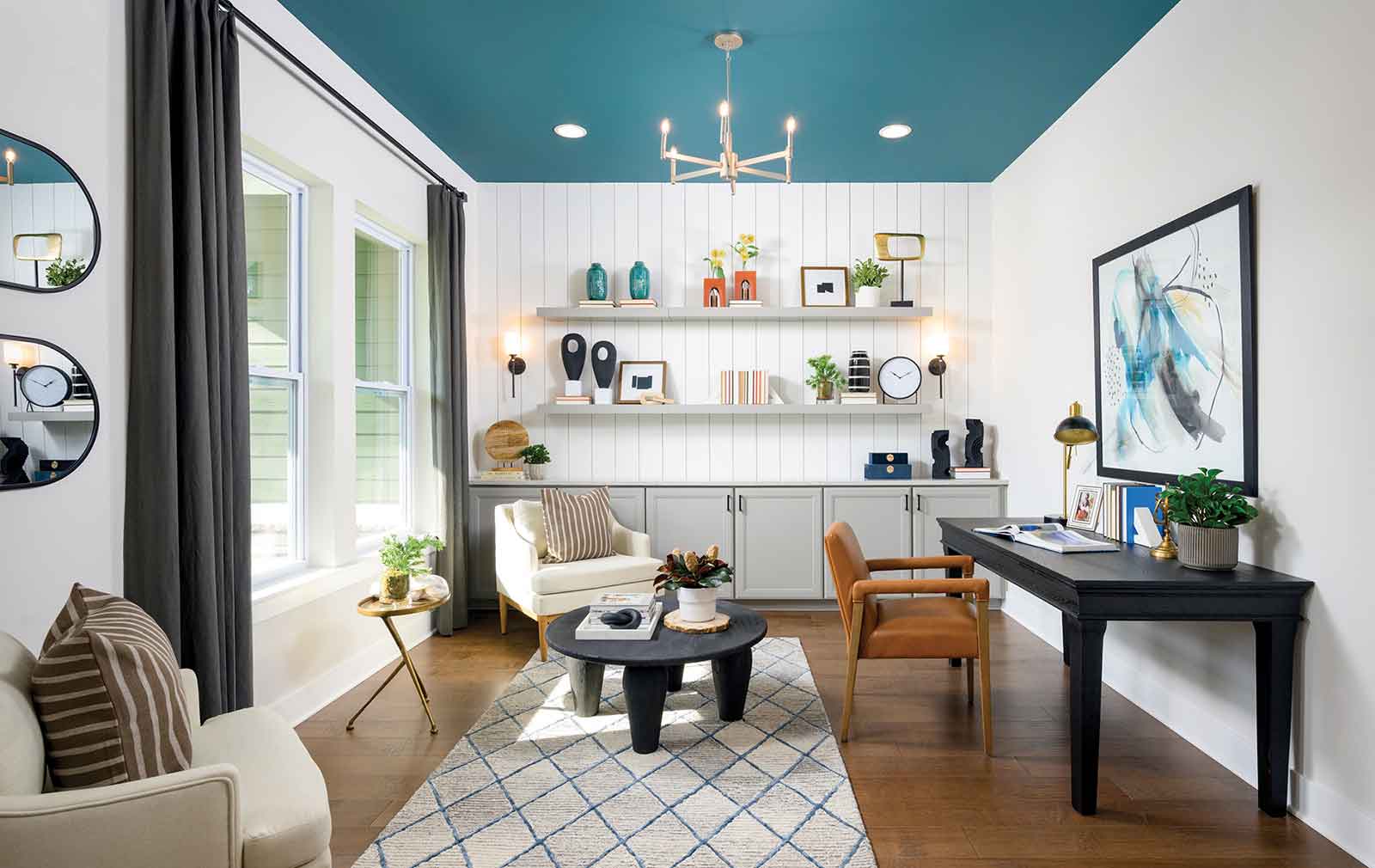 An open-concept office space with white walls, a blue ceiling, built-in shelves and cabinets at the back of the room, hardwood floors, two upholstered chairs facing a center table, and a wood chair pushed up to a black desk opposite the large front windows.