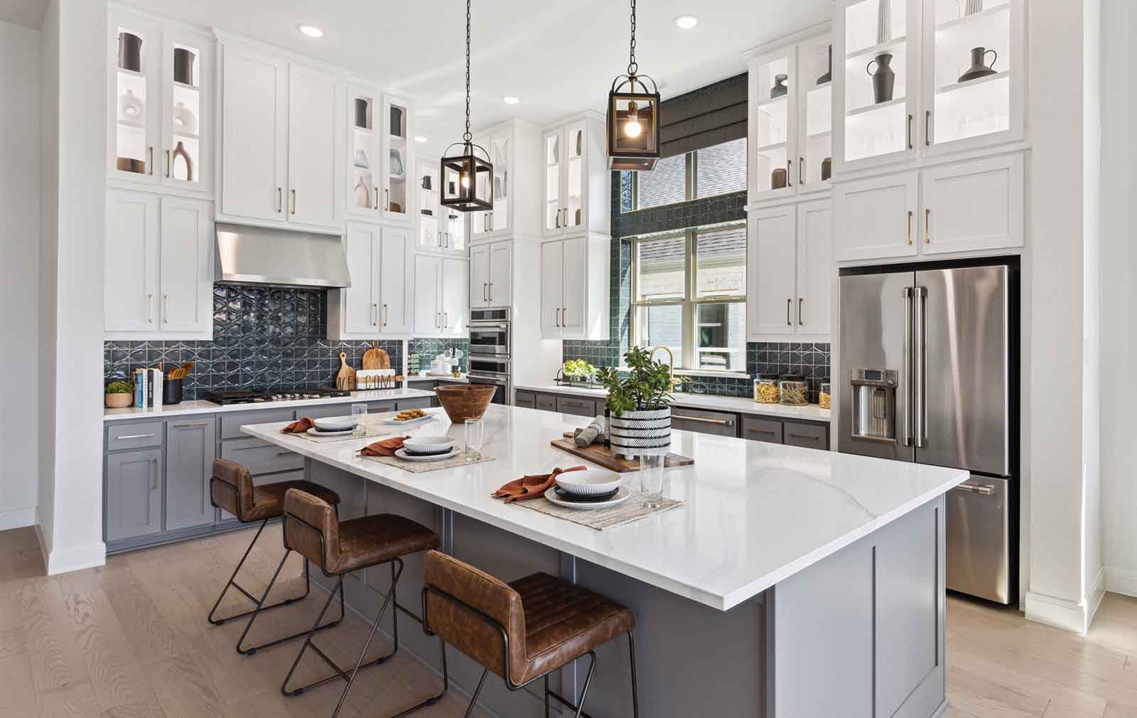 A model home’s kitchen with luxury wood-look plank flooring, leather upholstered stool seating pushed up to the white quartz-topped center island, a rich blue backsplash above light gray lower cabionets and clean, white upper cabinets that extend to the ceiling, including glass-fronted upper cabinets back-lit and displaying a variety of decorative pottery pieces.