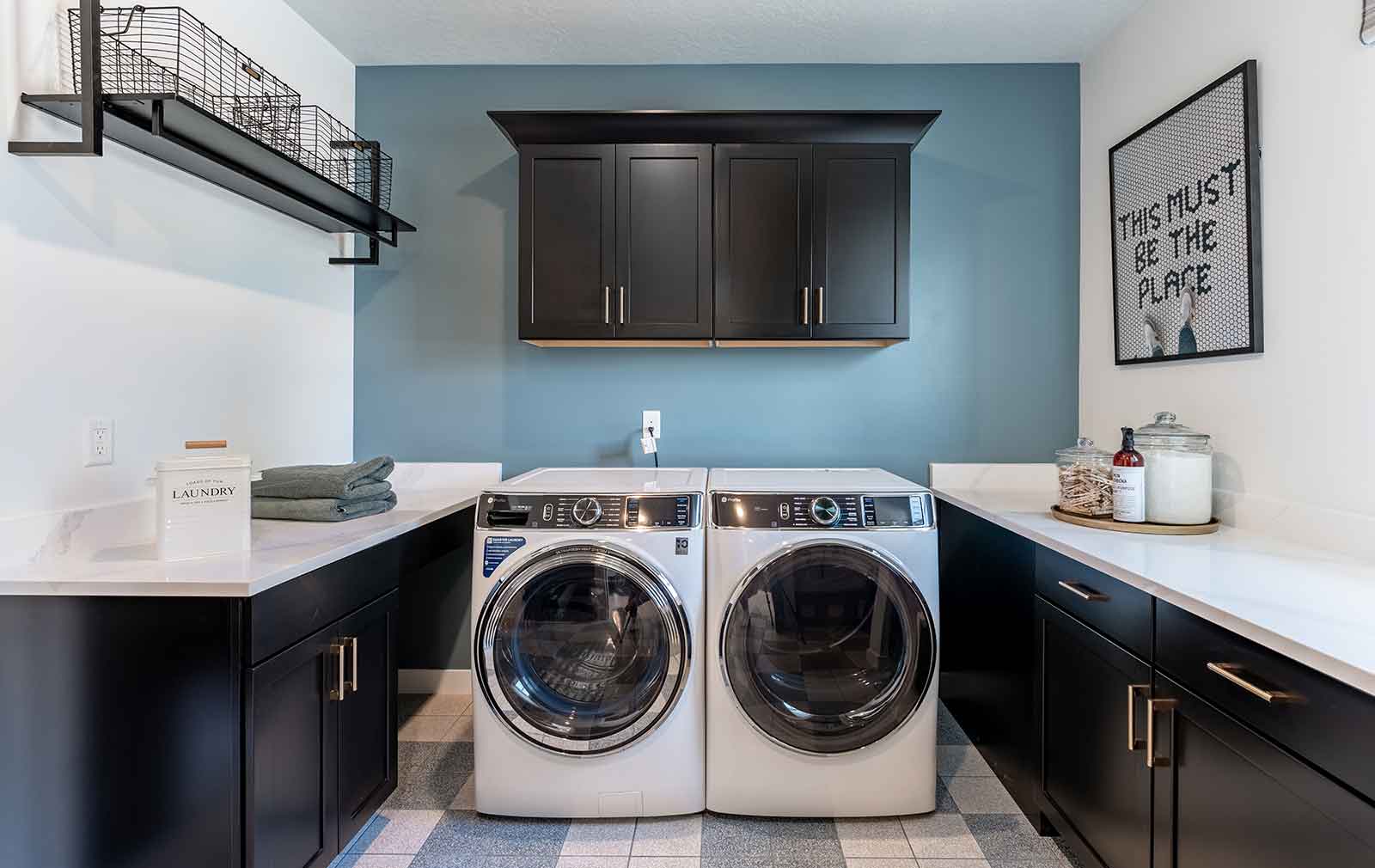 Blue and white checkerboard-pattern tiles support the classy black cabinetry with brass fixtures and white countertops, where retro laundry canisters and glass jars filled with powdered detergent give a classic flair and frame the sparkling clean front-load washer and dryer at the center of the frame.