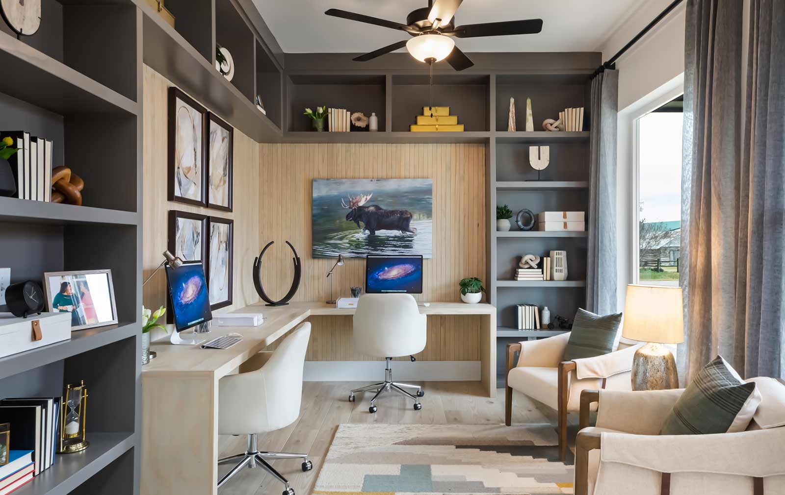 A model home’s study with two white sitting chairs and gray built-in shelving containing various décor items surrounding a white desk and hanging nature artwork.