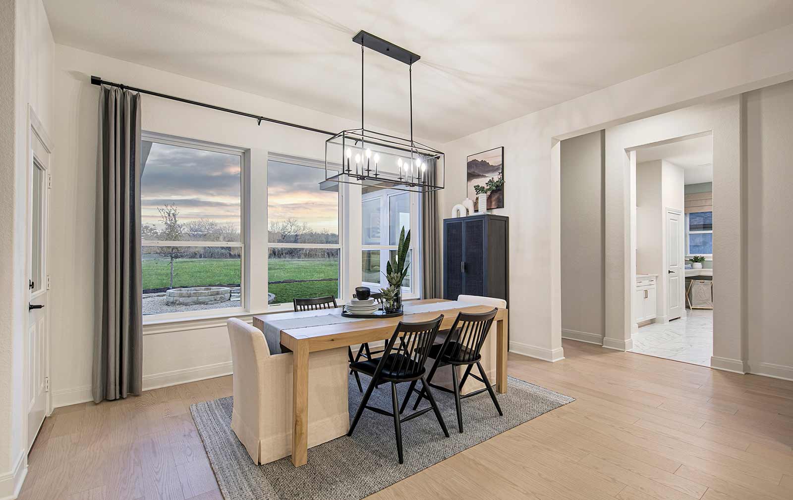 An expansive dining area with multiple large windows, light wood-like flooring, a small wooden table with three black wooden chairs and two cushioned chairs situated on a black and white area rug, a modern chandelier, a dark-colored armoire topped with ceramic decor, and artwork of a scenic mountain landscape hanging on the wall.