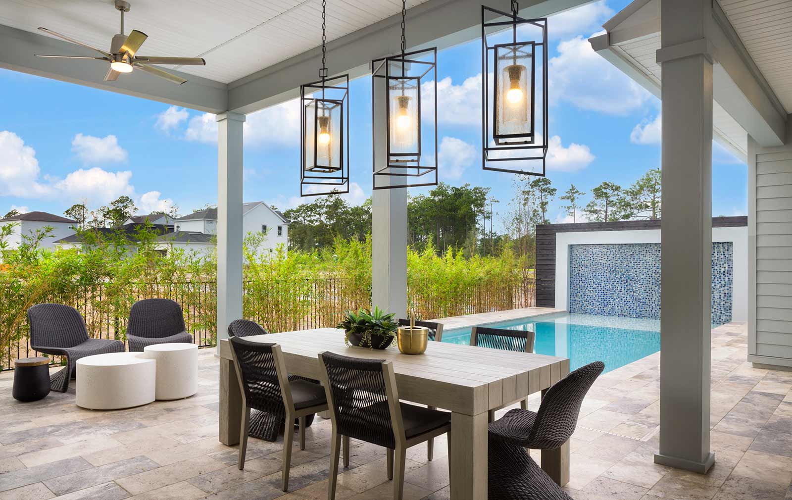 A model home’s covered patio with an outdoor dining table and lounge chairs in various gray tones and geometric light fixtures overlooks a swimming pool.