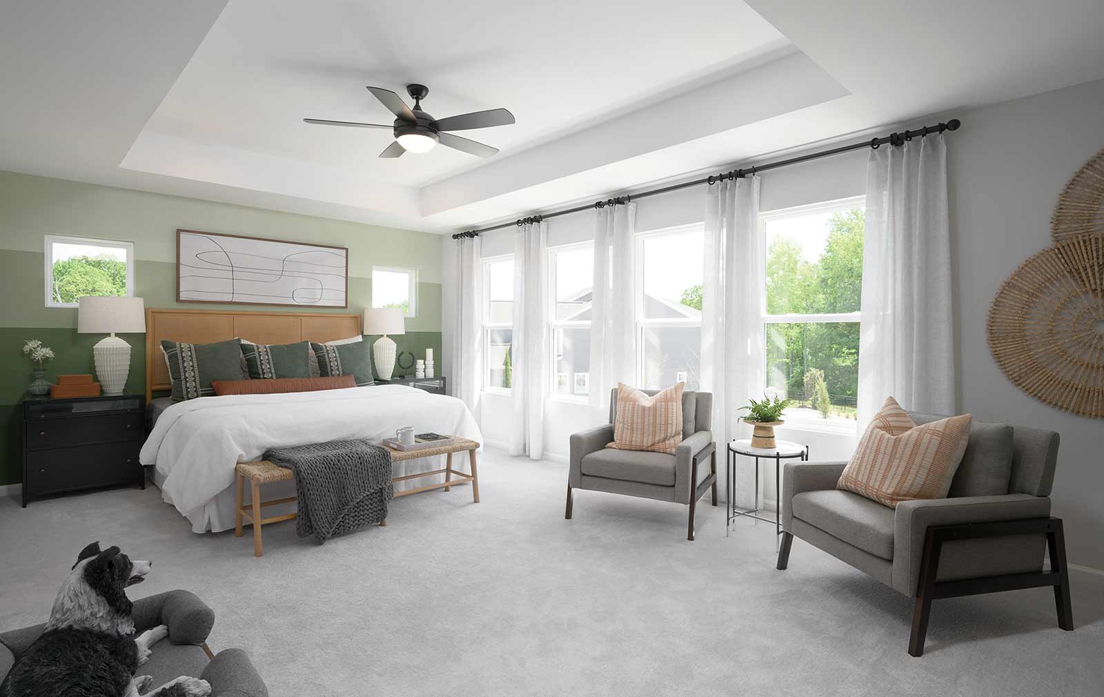 A model home’s Owner’s Retreat featuring white carpeting, a green-striped accent bedwall, wood and rattan décor, two gray sitting chairs, a row of windows, and a tray ceiling.