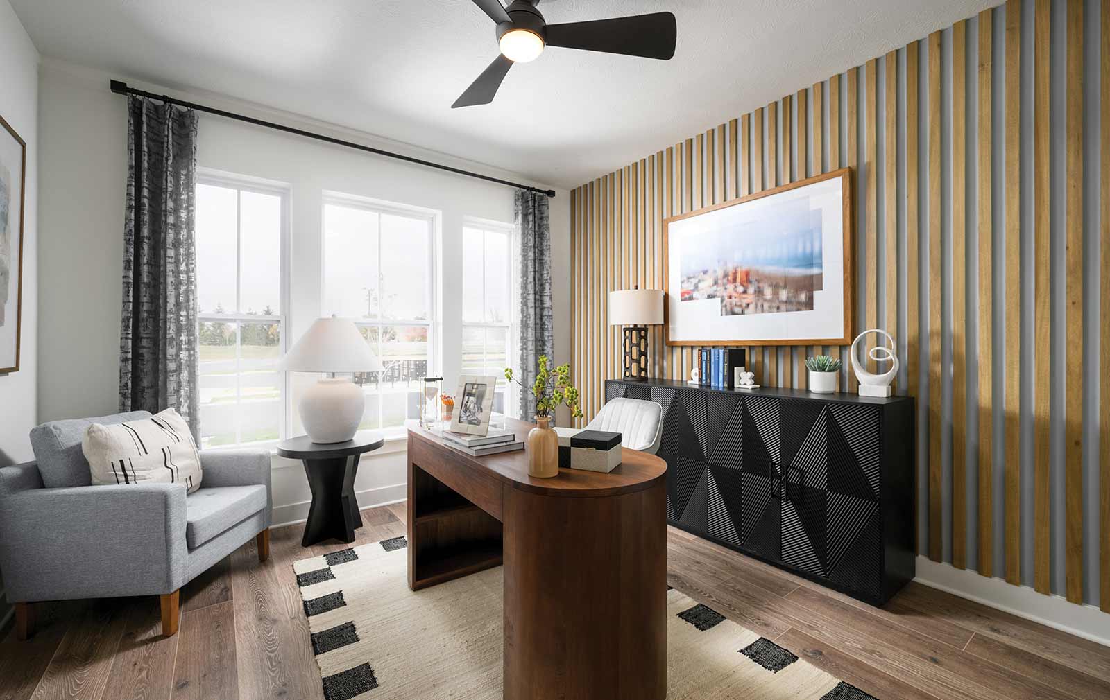 A study within a model home featuring geometric-style décor, a tan and gray paneled accent wall, a black sideboard, a black and white area rug, a white lamp, and a gray sitting chair with a black and white pillow.