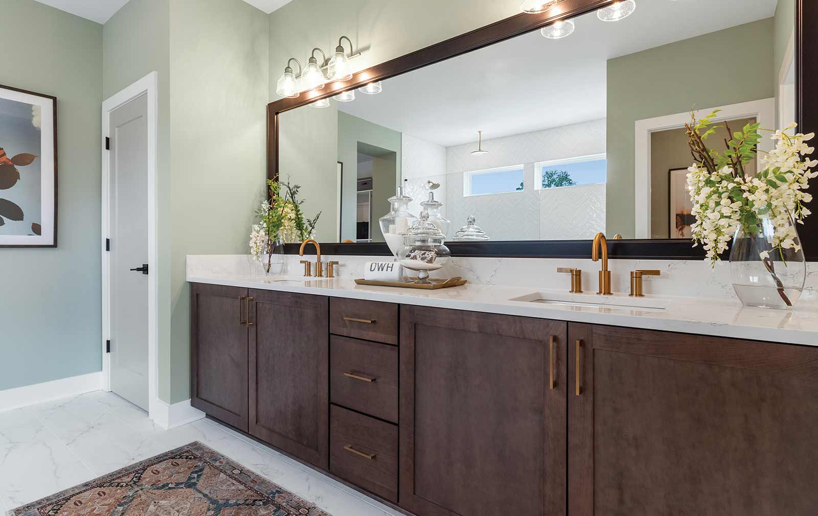 A model home’s Owner’s Bath with dark stained cabinets beneath dual sinks featuring gold finishes and a dark-framed mirror.